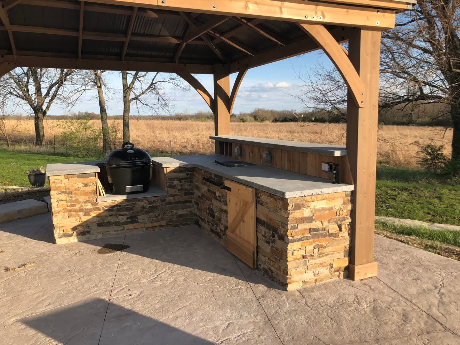 L-shaped outdoor kitchen with kamado grill and stone veneer under pergola